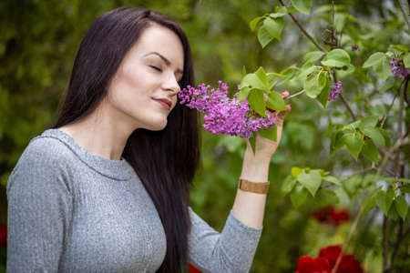 Young woman in gray dress smelling a lilac flower in summer