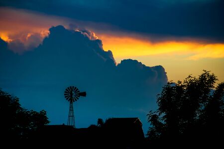 landscape with sky laden with storm clouds with yellow and blue orange colors and windmill silhouetteの写真素材