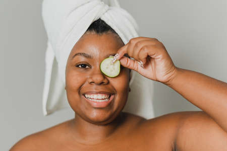Portrait of a happy young black woman with towel taking care of her skin with a natural cucumberの写真素材