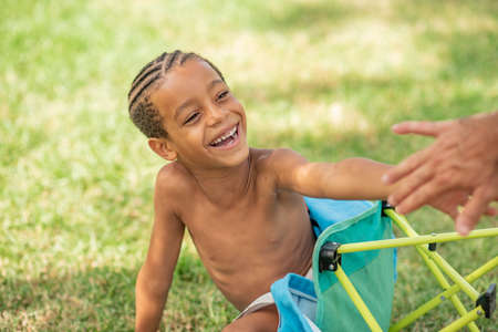 Delighted ethnic kid sitting on grassy lawn and playing with anonymous parent