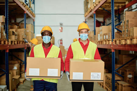 Multiracial warehouse workers holding delivery boxes while wearing safety mask for coronavirus...