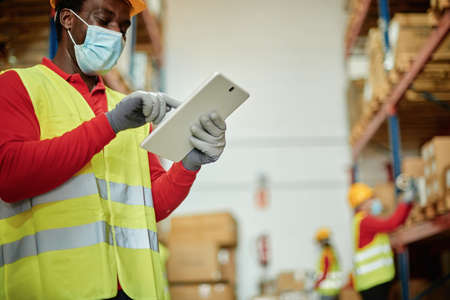 Afro male worker using a tablet checking the transport of a box in a warehouse wearing a safety mask...
