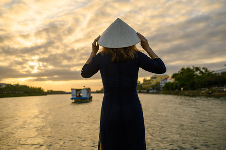 Back view of unrecognizable woman in Asian conical hat and long dress standing near river with rippling water and enjoying sunsetの写真素材