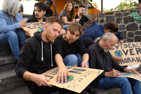 Group of focused diverse protesters creating carton placards while sitting on stairs during preparation for strikeの写真素材