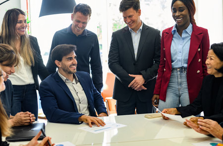 Team of glad multiracial coworkers in formal outfits standing at table with papers and discussing...