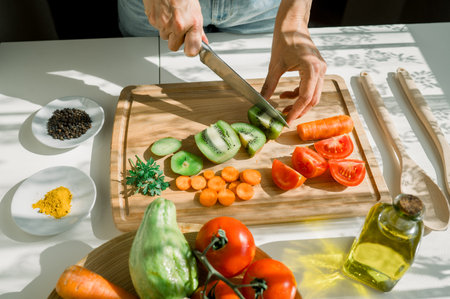 High angle of crop unrecognizable female cook cutting fresh kiwi on wooden chopping board while preparing salad with vegetables and fruits in kitchen on sunny dayの写真素材