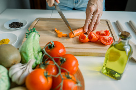 High angle of crop anonymous woman slicing fresh carrot on cutting board while preparing salad with tomatoes and parsley in kitchenの写真素材