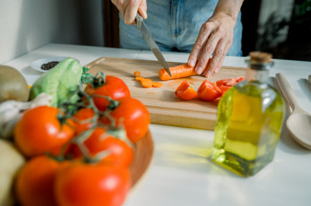 Crop anonymous housewife cutting carrot in kitchen while preparing healthy salad with tomatoes and herbs during cooking at homeの写真素材
