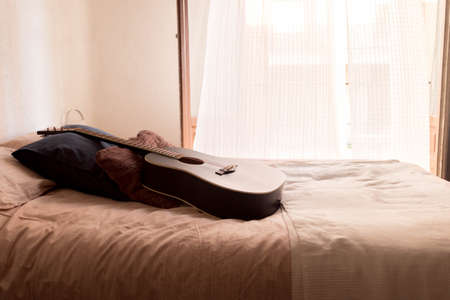 Black acoustic guitar on a bed a sunny day in a beautiful roomの写真素材
