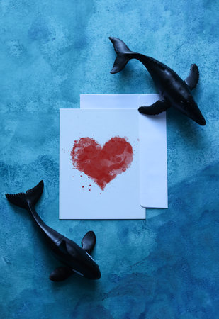 a still life with a card with a watercolor heart framed by toy whales on a blue watercolor backgroundの写真素材