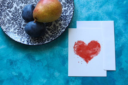 a still life with a plate with a floral pattern, ripe pear, plums and a paper card with a watercolor heart on a blue watercolor backgroundの写真素材