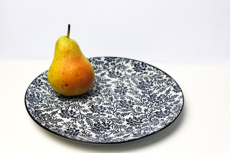 a bright yellow ripe pear on a plate with a floral pattern against a white backgroundの写真素材