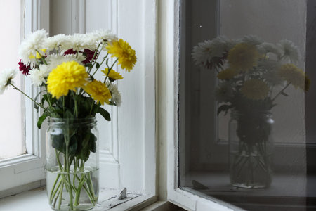 Bouquet of flowers in a glass jar placed on a windowsill, captured in soft natural light. The flowers are reflected in the windowの写真素材