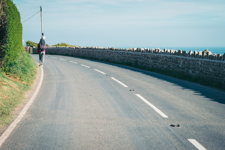 Female tourist, hichhiker, backpacker walking on the road to a distance in Rhossili, Walesの写真素材