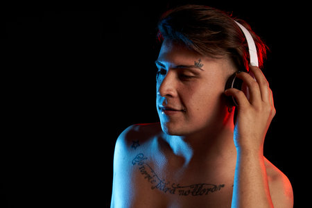 portrait latin man with headphones listening to music with tattoo "sonrie para no llorar" on black background and color lights copy spaceの写真素材