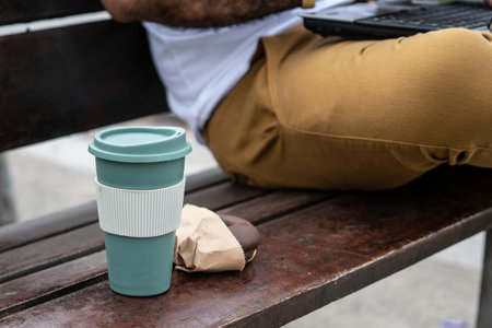 donut and mug of coffee on plaza bench, behind man's leg with laptop