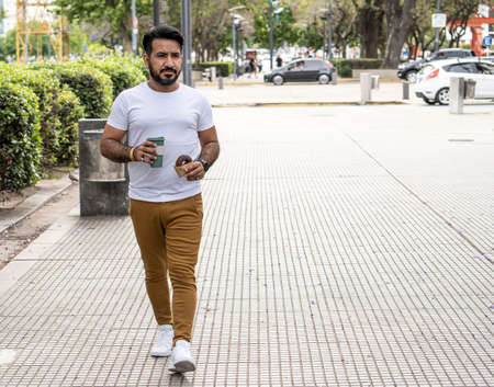 latin man walking in urban environment with mug of coffee and donutの写真素材