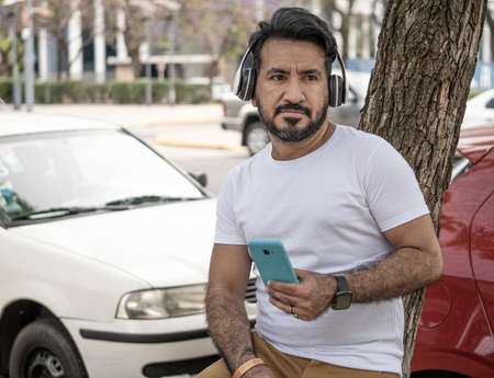 latin man with beard leaning against a tree in the city with cellular phone listening to musicの写真素材