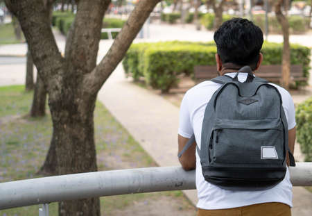 back shot of male tourist student leaning on a railing with a backpack looking into a park