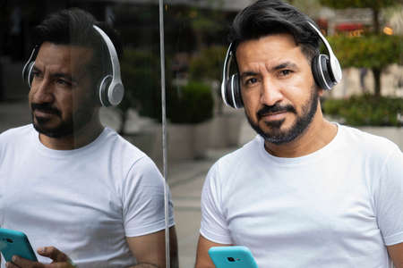 latin man with beard reflected in the glass of a building with cell phone and headphonesの写真素材