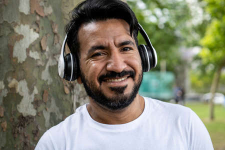 portrait Latin bearded man with headphones smiling at camera and listening to music in parkの写真素材