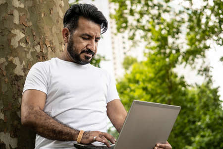 Latino man leaning on a tree with a laptopの写真素材