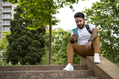 latino man sitting on the stairs of a park using his cell phoneの写真素材