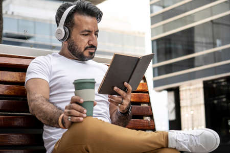 Latin man with beard sitting on a bench reading a book with a mug of coffee in an urban environment.