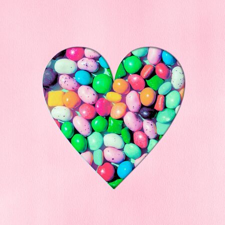 Heart shape filled with small colourful candies carved out of soft pink colored paper background....
