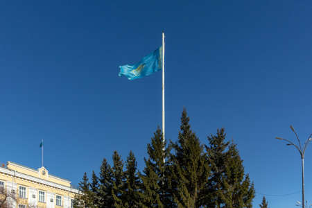 The flag of Kazakhstan hangs on a flagpole near the building behind the treesの写真素材