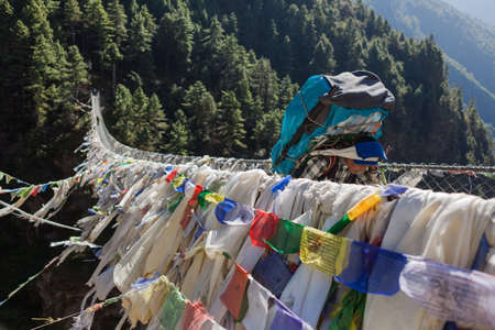 EVEREST BASE CAMP TREK/NEPAL - OCTOBER 19, 2015: Suspension bridge over the gorge in Sagarmatha National Park in Himalayas. Porter carrying big expedition bag over suspension bridge with prayer flags.のeditorial素材