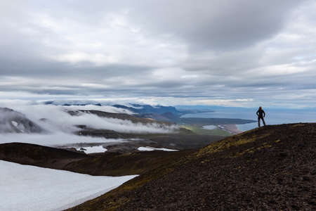 Solitude landscape in Iceland mountains. Single traveler standing on the hill and looking to the distant hills covered with white clouds and ocean. Beautiful mountains solitude landscape.の写真素材