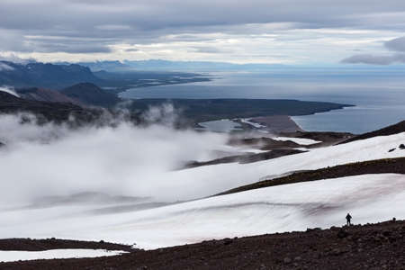 Solitude landscape in Iceland mountains. Single traveler standing on the slope of volcano looking to the distant sea shore. Beautiful mountains solitude landscape.の写真素材