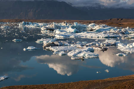 Scattered melting icebergs near Jokulsarlon glacier lagoon shore. Clouds reflecting in the water. Global warming and climate change concept with melting ice. Vatnajokull glacier, Iceland.の写真素材