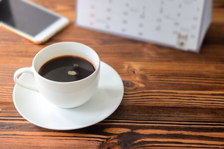 Coffee in a white cup with calendar and smart phone on wooden desk in officeの写真素材
