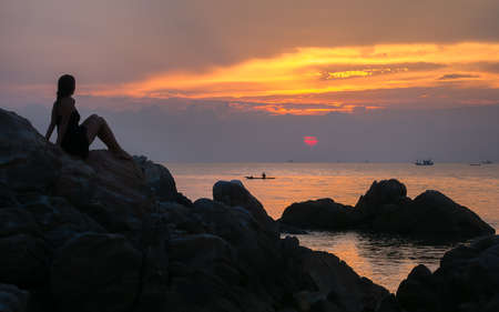 Silhouette of the girl watching the sea sunset and small boats sitting on the rocks.の写真素材