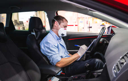 A young caucasian technician doing a security inspection inside a vehicle protected with a mask and gloves to prevent the spread of virusの写真素材