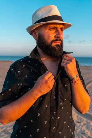 Portrait of a man with a beard and a white hat, putting on his shirt, at sunset on a beach