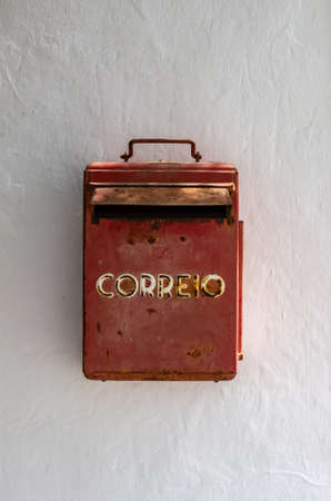Old and rusty red mailbox in a white wall between a tree and a door in a traditional Canary house....