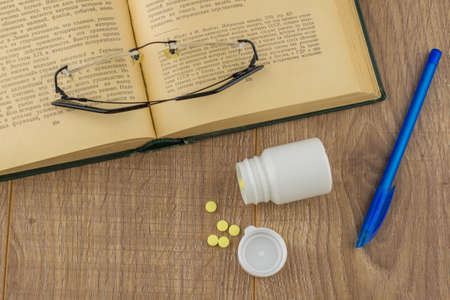 drowsiness and malaise at work: a box of pills to combat the drowsiness and weakness, glasses, open book, blue pen - top viewの写真素材