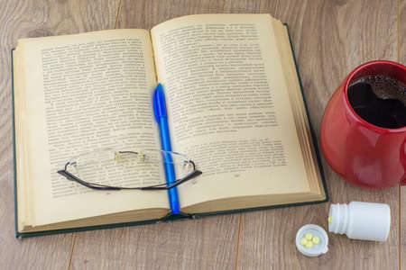 Sleepiness and insomnia: caffeine pills near the pill box, old book, glasses and red large mug of strong coffee on the table, blue pen on the bookの写真素材