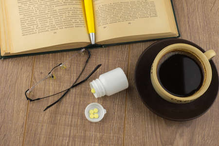 Drowsiness: caffeine pills near the pill box, old book, glasses and mug of strong coffee on the wooden table, yellow ball-point pen on the book closeの写真素材