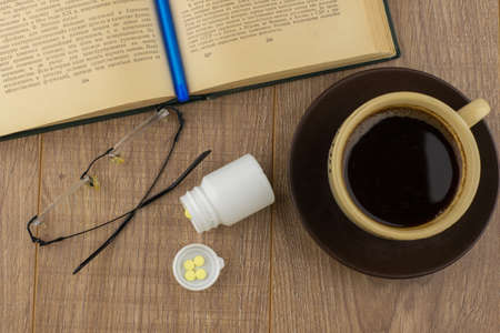 Drowsiness: caffeine pills near the pill box, old book, glasses and mug of strong coffee on the wooden Desk, blue pen on the book closeの写真素材