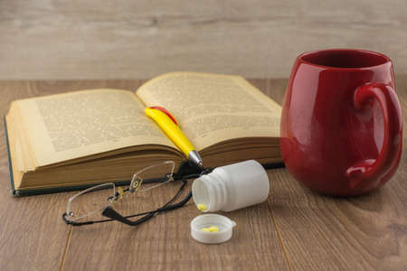 Drowsiness: caffeine pills near the pill box, old book, glasses, and a red mug of strong coffee on the wooden table, yellow ball-point pen on the book, front viewの写真素材
