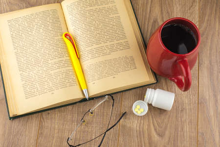 Drowsiness: caffeine pills near the pill box, old book, glasses, and a red mug of strong coffee on the wooden table, yellow ball-point pen on the book, the view from the topの写真素材