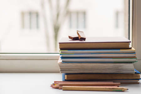 A stack of old different books and several colored pencils lie on the windowsill, a couple of pieces of chocolate lying on the booksの写真素材