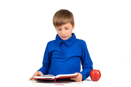 boy schoolboy in a blue t-shirt is preparing for school, reading something in a notebook, next to the table is a red appleの写真素材