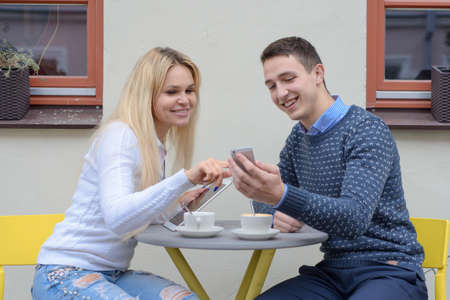 a young man and a blonde girl with long hair drink coffee or tea next to a cafe at a table, a guy shows something to a girl on his phone, they laughの写真素材