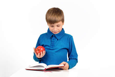 boy schoolboy in a blue t-shirt is preparing for school, reading something in a notebook, holding a red appleの写真素材