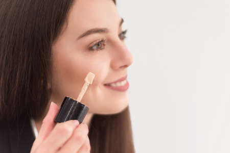 girl, make-up artist, with long dark hair in a business suit, applies a concealer on the face on a white backgroundの写真素材